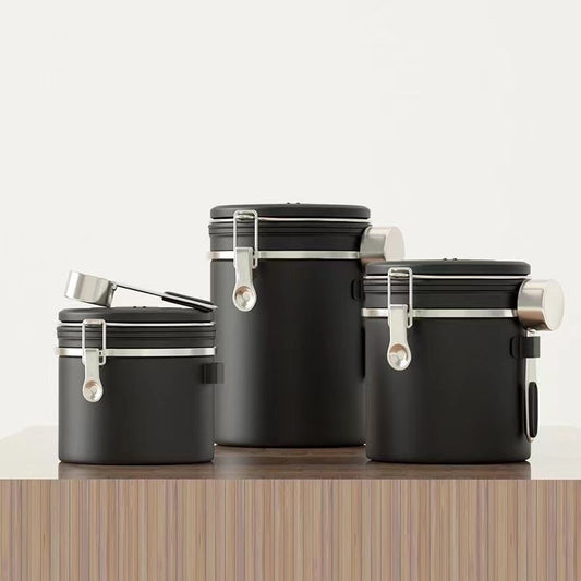 A set of three black airtight coffee canisters with a matching stainless steel scoop on a wooden table. The canisters feature a modern design with a latch and a date tracker on the lid. Perfect for keeping coffee beans fresh." Shop now!
