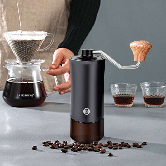 Handheld coffee grinder with coffee beans and a coffee brewing setup on a gray surface.