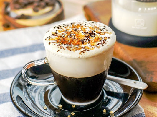 Espresso shot with whipped cream and toppings on a saucer, with a blurred background of a kitchen setting.