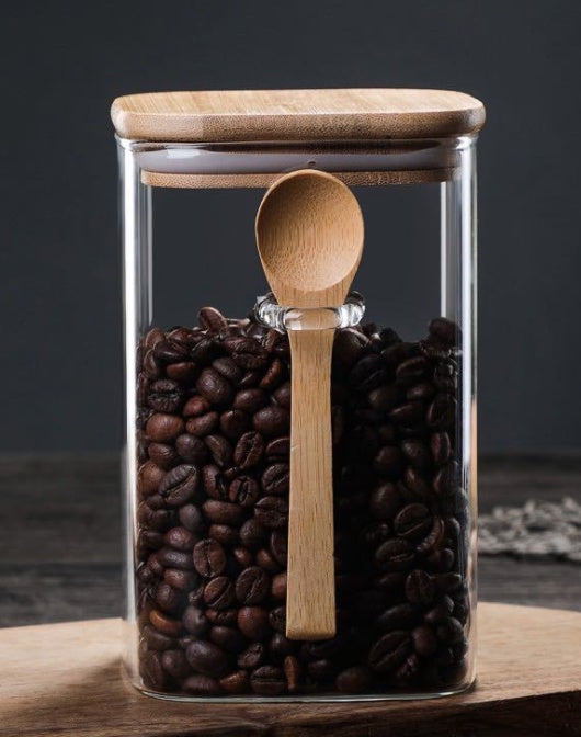 A square glass canister filled with roasted coffee beans. The canister has an airtight bamboo lid and a wooden spoon held in a built-in glass loop on the side. The image is on a dark wood surface.