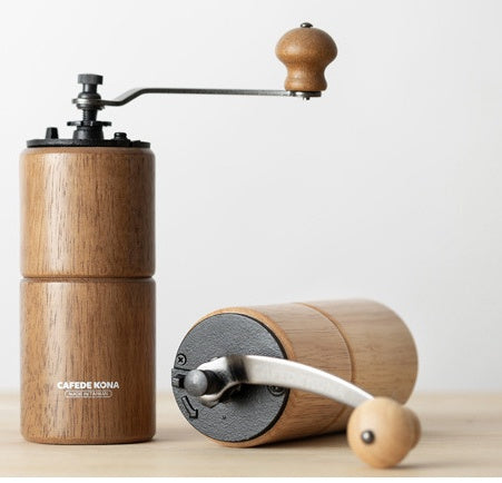 Two CAFÉDE KONA manual coffee grinders, one light wood upright, one dark wood lying down, on wood surface.