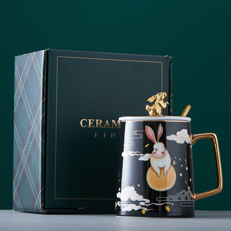 Ceramic mug with artistic design and gold handle next to a gift box on a dark green background