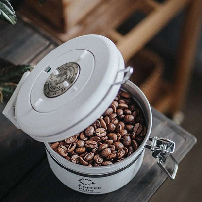 A grey Coffee Plus Breathing Canister, open with a 7g coffee scoop resting on a pile of fresh coffee beans. Another canister is visible in the background. Show now