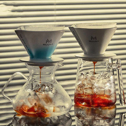 A hand places a paper filter into a Muvna Hoshikawa ceramic pour-over dripper in Hoshikawa Blue, highlighting the brewing process. The image showcases the beautiful two-tone glaze and the complete set with the glass carafe. Shop Now!