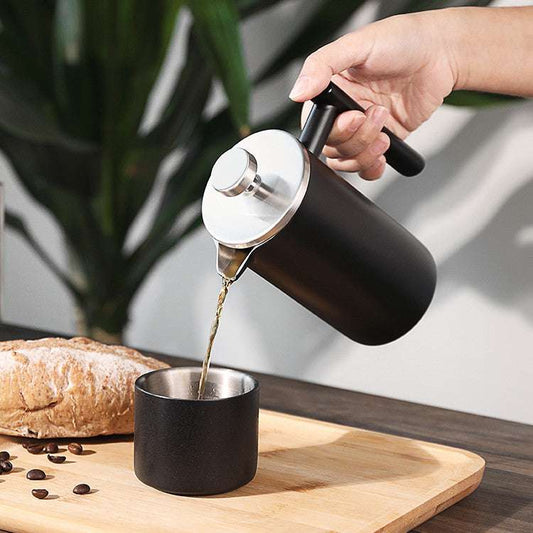 Person pouring coffee from a black French press into a black mug on a wooden cutting board with bread and coffee beans. Shop now! Free shipping