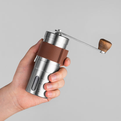 Hand holding compact stainless steel manual coffee grinder with brown silicone band, showcasing portability and ergonomics.