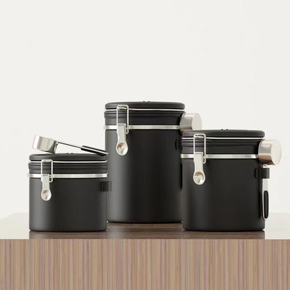 A set of three black airtight coffee canisters with a matching stainless steel scoop on a wooden table. The canisters feature a modern design with a latch and a date tracker on the lid. Perfect for keeping coffee beans fresh." Shop now!