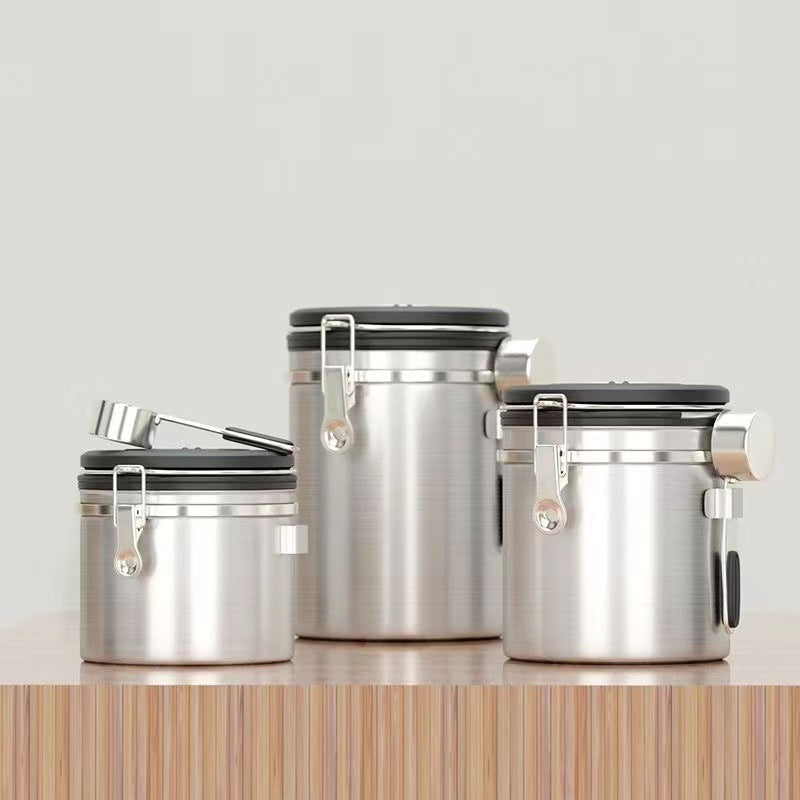 A set of three silver airtight coffee canisters with a matching stainless steel scoop on a wooden table. The canisters feature a modern design with a latch and a date tracker on the lid. Perfect for keeping coffee beans fresh." Shop now!