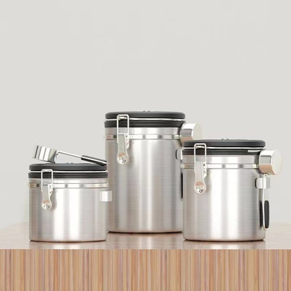 A set of three silver airtight coffee canisters with a matching stainless steel scoop on a wooden table. The canisters feature a modern design with a latch and a date tracker on the lid. Perfect for keeping coffee beans fresh." Shop now!