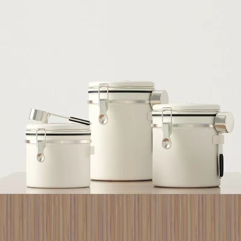 A set of three white airtight coffee canisters with a matching stainless steel scoop on a wooden table. The canisters feature a modern design with a latch and a date tracker on the lid. Perfect for keeping coffee beans fresh." Shop now!