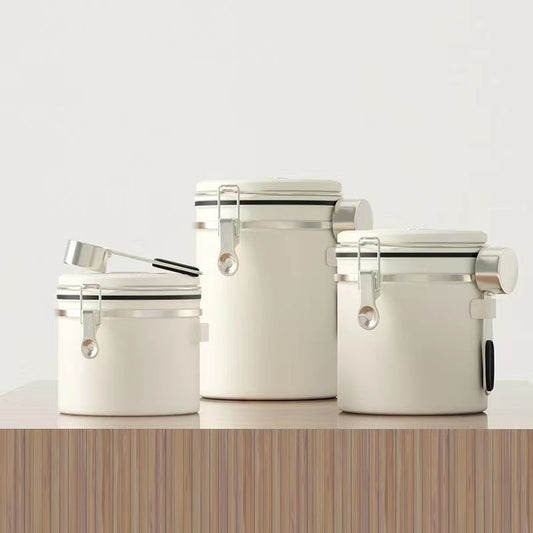 A set of three white airtight coffee canisters with a matching stainless steel scoop on a wooden table. The canisters feature a modern design with a latch and a date tracker on the lid. Perfect for keeping coffee beans fresh." Shop now!