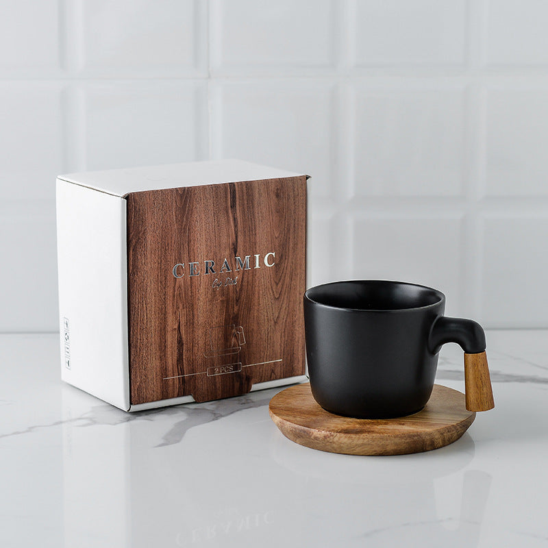 Black ceramic mug with wooden handle on a coaster next to a branded wooden box on a light gray surface. Gift box