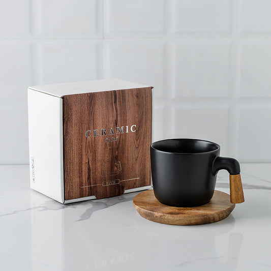 Black ceramic mug with wooden handle on a coaster next to a branded wooden box on a light gray surface. Gift box