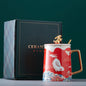 Ceramic mug with decorative design and gold handle next to a black gift box on a green background