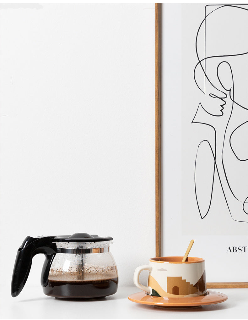 Coffee maker and cup with spoon on a table next to a framed abstract art piece.