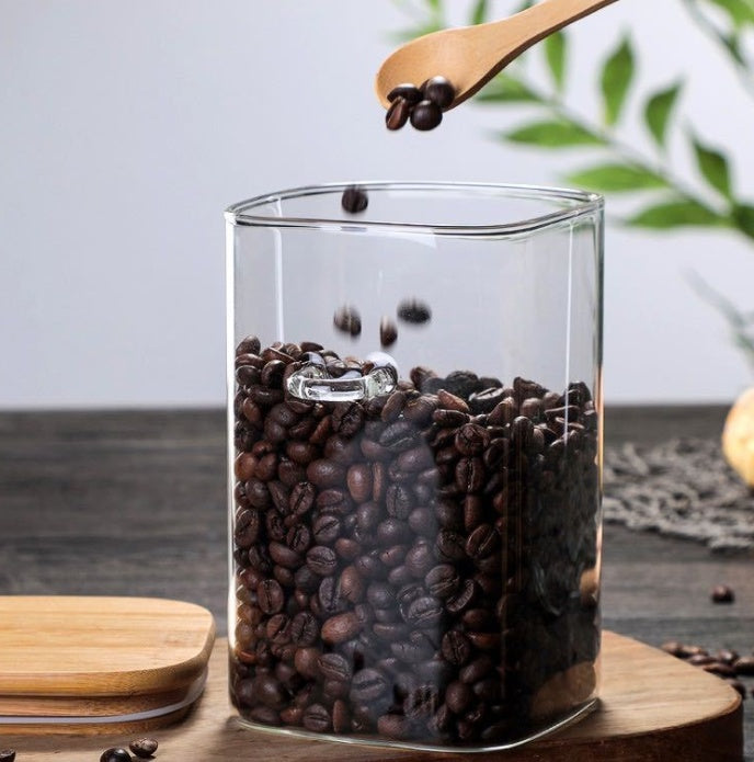 A person is pouring fresh roasted coffee beans into a clear square glass canister. The canister has a built-in loop for a wooden spoon and is partially filled with beans. The background is a wooden table. Shop now
