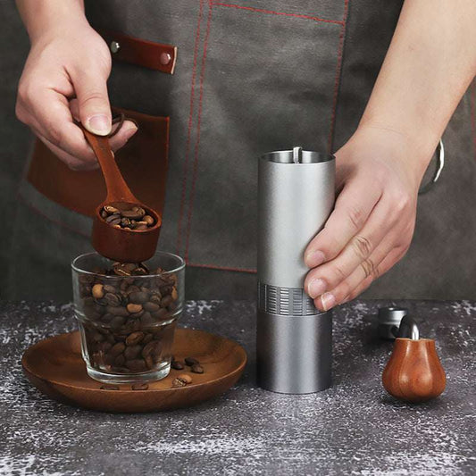 Person loading silver manual coffee grinder with whole coffee beans for fresh grinding.