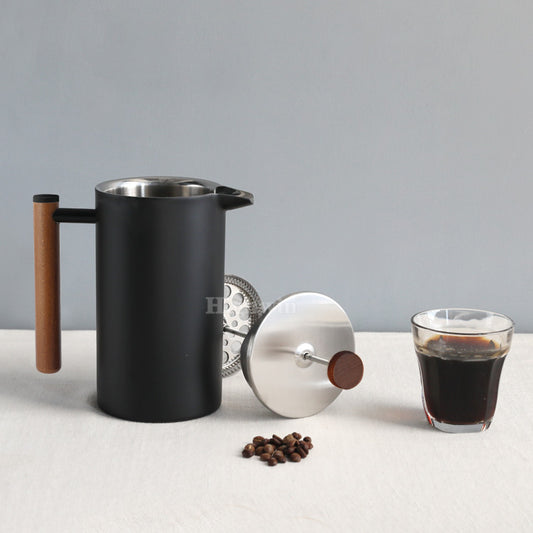 Black coffee press with wooden handle, glass of coffee, and coffee beans on a light surface.