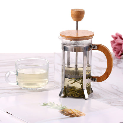 Glass French press with wooden lid and handle, filled with tea leaves, next to a glass mug with tea on a white surface. Free shipping + Shop now