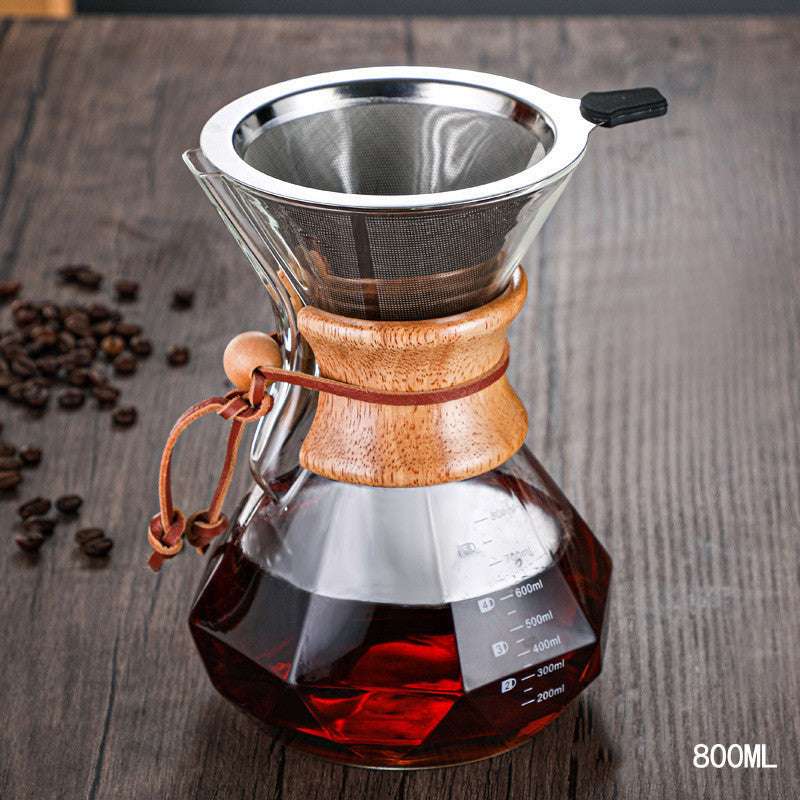 Clear glass coffee maker with wooden collar and metal filter on a wooden surface with coffee beans.