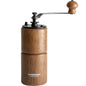 Two CAFÉDE KONA manual coffee grinders, one light wood upright, one dark wood lying down, on wood surface.