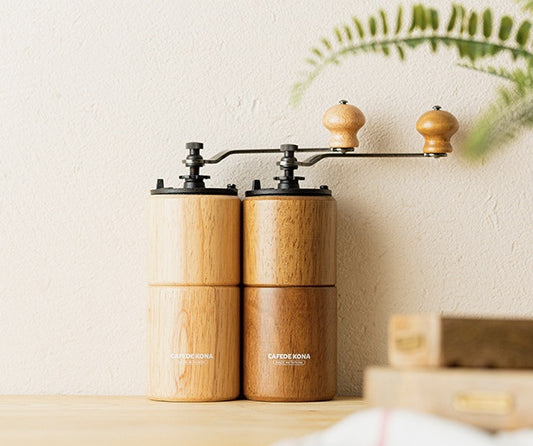 CAFÉDE KONA manual coffee grinders light and dark wood side by side on table, home coffee brewing.